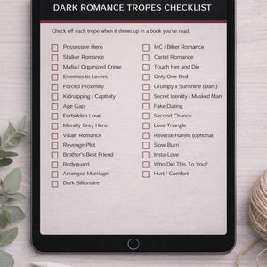 K&ouml;nnte beinhalten: Ein Tablet-Computer zeigt eine Checkliste mit dem Titel &bdquo;Dark Romance Tropes Checklist&ldquo;. Die Liste enth&auml;lt Roman-Tropen wie &bdquo;Besitzergreifender Held&ldquo;, &bdquo;Mafia&ldquo; und &bdquo;Arrangierte Ehe&ldquo;. Ein Stift, eine goldene B&uuml;roklammer und ein Garnkn&auml;uel liegen auf dem Tisch.
