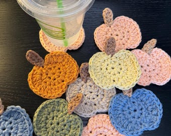 May include: A collection of small, round, crocheted apple-shaped coasters in various pastel and muted colors including peach, yellow, blue, olive green, and grey, each featuring a small brown stem, arranged in a cluster with one under a clear cup.