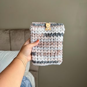 May include: A rectangular crochet pouch featuring a variegated yarn pattern in shades of cream, light brown, and dark gray. The pouch is secured at the top center with a small, light-colored wooden button shaped like a teddy bear face.