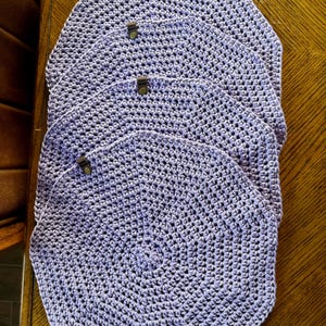 May include: Set of four lavender crocheted placemats with a circular design. Each placemat has a brown leather tab.
