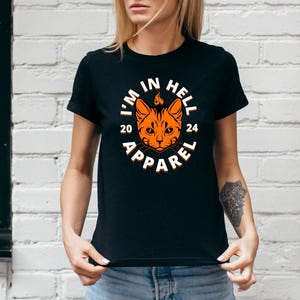May include: Black crew neck t-shirt with a circular graphic featuring an orange cat face with flames above its head. The text "I'M IN HELL 2024 APPAREL" is printed around the cat's face in white.
