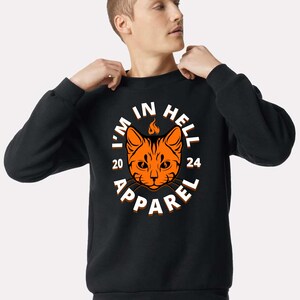 May include: Black crew neck sweatshirt with a graphic featuring an orange cat face with a flame above its head. The text "I'M IN HELL 2024 APPAREL" is printed in white around the cat design. The sweatshirt is a solid color.