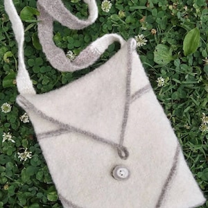 May include: A white knitted crossbody bag with a gray trim and a single button closure. The bag is lying on a bed of green clover.