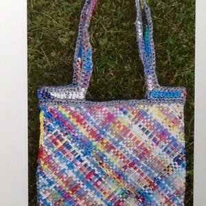 May include: A woven tote bag with a colorful, diagonal pattern in shades of blue, red, yellow, and white. The bag has gray handles and trim. Text at the top reads "Folded Plarn Bag with liner" and at the bottom "By Laura Haines."