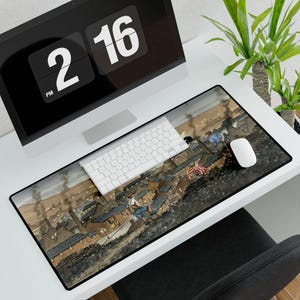 May include: A rectangular desk mat featuring an illustration of a town with people on rooftops, smoke stacks, and a cloudy sky. A white keyboard and mouse sit on the mat, with a computer monitor and a plant in the background.