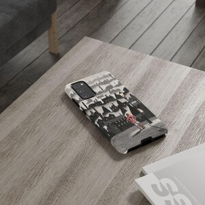 May include: A phone case with a black and white cityscape illustration, including a person on a cobbled street. The case has a white top and black sides. It rests on a wooden table.