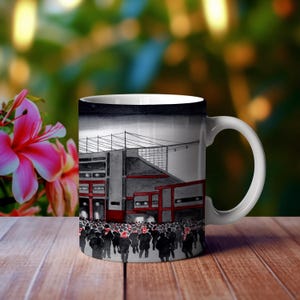 May include: A white ceramic mug featuring a black and white illustration of a stadium with a crowd of people in front of it. The stadium is red and grey.
