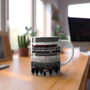 Peut inclure: Une tasse en céramique blanche avec une illustration en noir et blanc d'une foule de personnes devant un stade. Le stade porte l'inscription "Manchester United Football Club" sur sa façade.