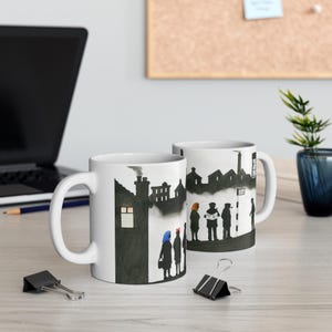 May include: White ceramic mug featuring a black and white illustration of a street scene with people walking. The illustration includes a chimney, houses, and a sign that reads "Stop".