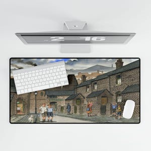 May include: A rectangular desk mat featuring a detailed watercolor painting of a village street scene with brick buildings, figures, and a dog. A white keyboard and mouse are on the mat. The time 2:16 is displayed on the monitor.