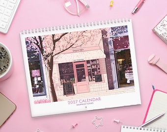 May include: A 2027 calendar titled Shoppront Scenes featuring an illustration of a storefront with a pink door, a striped awning, and cherry blossom trees. The text beginnings and new beginnings are printed above the shop entrance.
