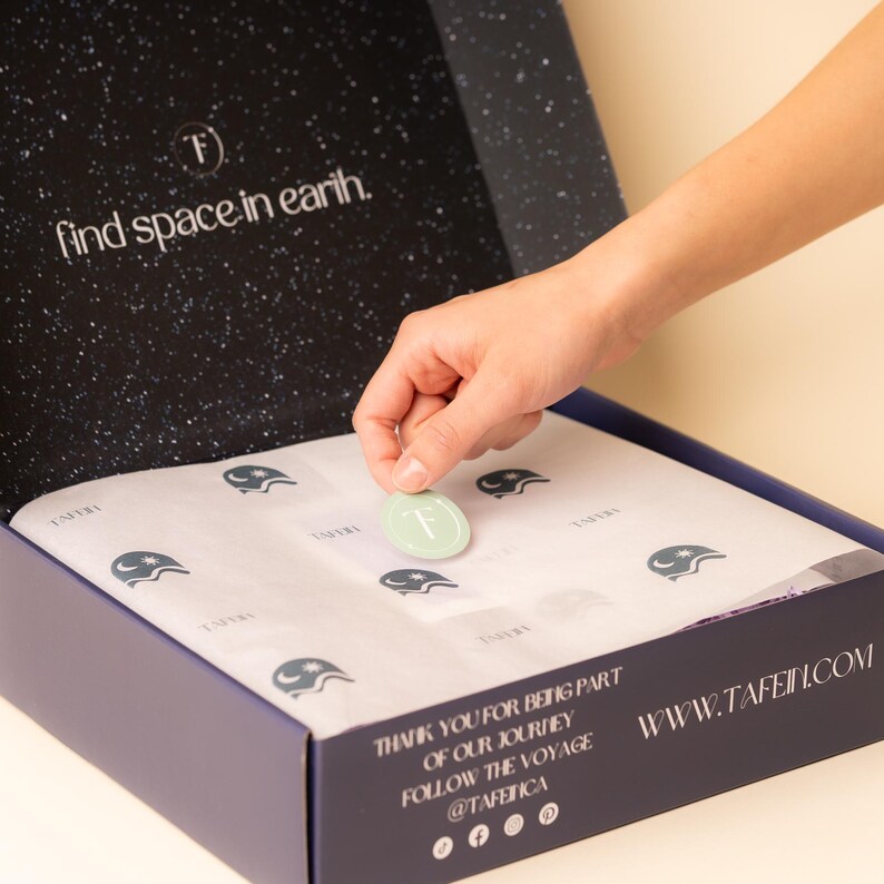 May include: A blue box with a white star pattern and the text "find space in earth."  The box is open and a hand is placing a sticker with the letter "F" on the inside of the box. The box also has the text "WWW.TAFE.COM" and "THANK YOU FOR BEING PART OF OUR JOURNEY FOLLOW THE VOYAGE @TAFETICA" on the side.