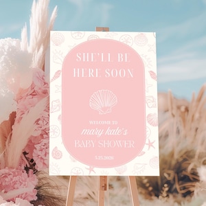 Könnte beinhalten: Ein rosa-cremefarbenes Babyparty-Schild auf einer Holzstaffelei. Auf dem Schild steht "She'll Be Here Soon" und "Welcome to Mary Kate's Baby Shower" mit einer Muschel-Grafik. Der Hintergrund zeigt rosa und cremefarbene Blumenarrangements.