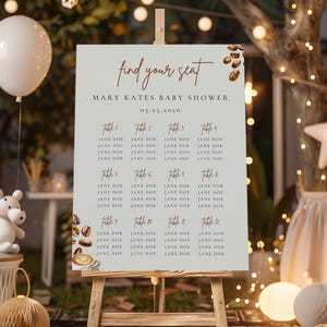 May include: A light beige seating chart on a wooden easel for a baby shower. The chart features the text "find your seat" and guest names organized by table numbers. Coffee bean illustrations and decorative elements surround the text.