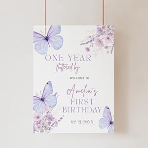 May include: A white sign with a butterfly and floral design, featuring the text "ONE YEAR Fluttered by WELCOME TO Amelia's FIRST BIRTHDAY". The sign has a light purple color scheme with butterflies and flowers.