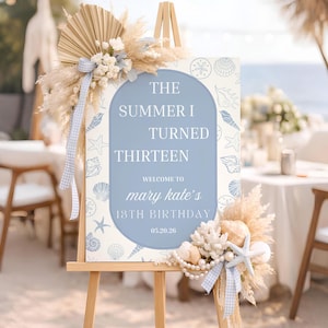 Puede incluir: Un caballete de madera exhibe un letrero con el texto "The Summer I Turned Thirteen" en un marco azul arqueado. El letrero está decorado con conchas marinas y estrellas de mar, y adornado con flores secas y cintas.