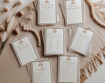 May include: A selection of baby shower game cards, featuring titles such as "Nursery Rhymes" and "Baby Bingo." The cards have a cream background with brown text and decorative borders. The image is styled with neutral-toned decor, including wooden beads and dried flowers.