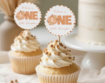 May include: Two cupcakes with white frosting and sprinkles, topped with round toppers that read "Olivia is ONE Little Pumpkin" with a pumpkin graphic. A cake and vase are in the background. The cupcakes sit on a white surface.