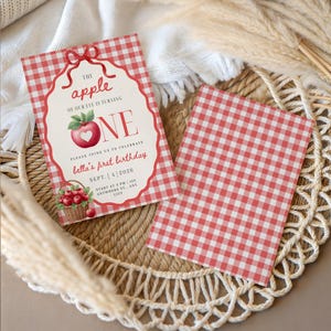 Op de afbeelding: Een rechthoekige verjaardagsuitnodiging met een rood-wit ruitpatroon, voorzien van een illustratie van een rode appel met een hartvormige uitsnede, een mand met appels en de tekst The apple of our eye is turning ONE, Bella's first birthday.