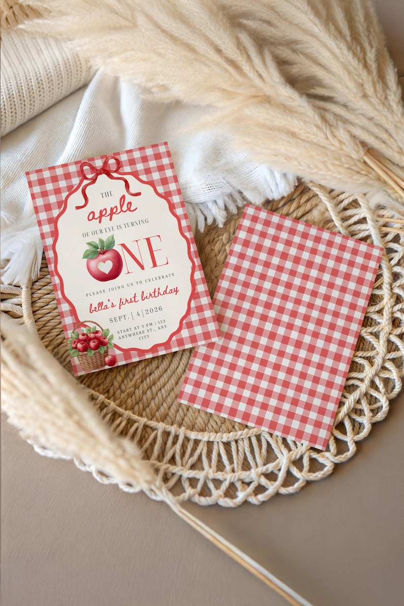 Op de afbeelding: Een rechthoekige verjaardagsuitnodiging met een rood-wit ruitpatroon, voorzien van een illustratie van een rode appel met een hartvormige uitsnede, een mand met appels en de tekst The apple of our eye is turning ONE, Bella's first birthday.