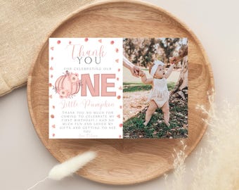 May include: A thank you card with a photo of a baby and the text "Thank You" and "One Little Pumpkin". The card has a pink and floral design, suitable for a first birthday celebration. The card is displayed on a wooden plate.