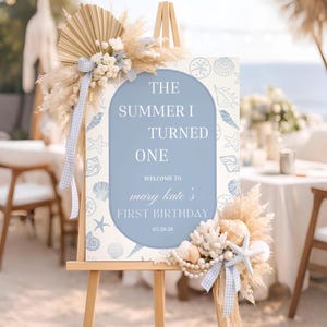 May include: A wooden easel holds a sign with the text "The Summer I Turned One" and "Mary Kate's First Birthday." The sign is decorated with seashells, starfish, and floral arrangements in blue, white, and beige. The event is a beach-themed first birthday party, with a coastal aesthetic.