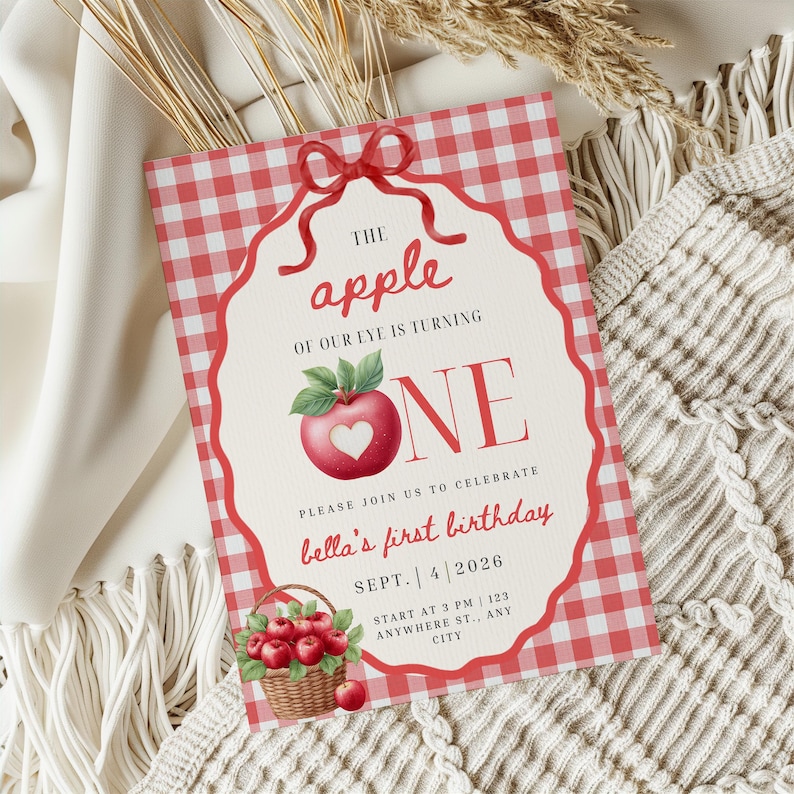 Op de afbeelding: Een rechthoekige verjaardagsuitnodiging met een rood-witte ruitjesrand, voorzien van een rode appel met een hartvormige uitsparing, een mand met appels en de tekst THE apple OF OUR EYE IS TURNING ONE, bella's first birthday, SEPT. 4 2026.