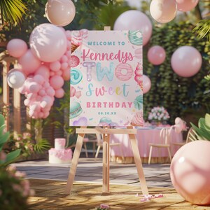 Puede incluir: Un caballete de madera exhibe un letrero que dice "Welcome to Kennedys Two Sweet Birthday" con ilustraciones coloridas de dulces. El letrero est&aacute; decorado con elementos rosas, azules y verdes, y est&aacute; rodeado de globos rosas.