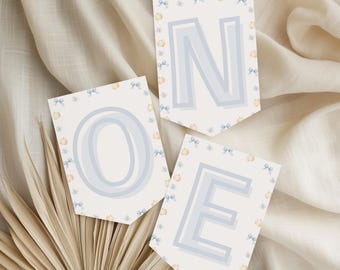 May include: Three banner flags spelling "ONE" in light blue, with a bow and pumpkin pattern border. The flags are on a cream-coloured background with a dried palm leaf in the lower left corner. The letters are outlined in a darker blue.