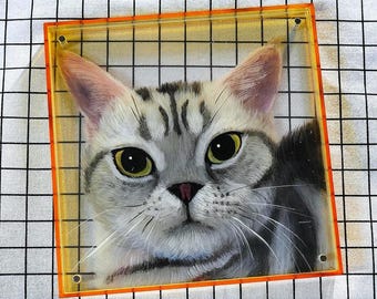 Op de afbeelding: Een close-up van een schilderij van een grijs-witte tabbykat met groene ogen. De kat kijkt recht naar de kijker. Het schilderij is ingelijst in een helder acryl frame.