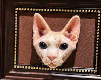 May include: A close-up of a felted sculpture of a hairless cat's head in a brown wooden frame. The cat has blue eyes and a pink nose.