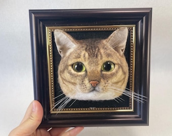 Puede incluir: Una cabeza de gato de fieltro de aguja enmarcada con pelaje marrón y blanco, ojos verdes y fondo negro.