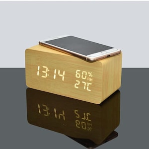 Könnte beinhalten: Ein helles digitales Wecker aus Holz mit einem Smartphone obenauf. Die Uhr zeigt Zeit, Luftfeuchtigkeit und Temperatur in weißen LED-Leuchten an. Die Uhrzeit ist 13:14. Die Luftfeuchtigkeit beträgt 60% und die Temperatur 27°C.