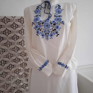 May include: White long-sleeved blouse with blue and black floral embroidery on the neckline and cuffs.