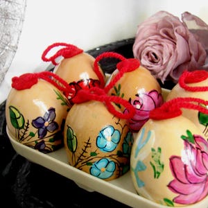 May include: Six hand-painted wooden eggs with floral designs in shades of blue, pink, purple, and red. Each egg has a red string attached to the top.