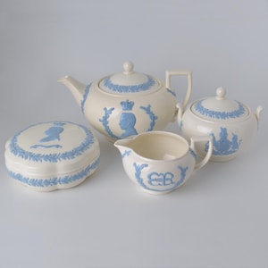 Puede incluir: Un juego de té color crema con diseños en relieve azules. El juego incluye una tetera, azucarero, lechera y una pequeña caja redonda. Cada pieza presenta un perfil de una persona con corona.
