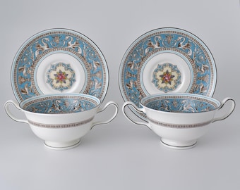 Può includere: Due tazze da tè bianche con manici e piattini abbinati. Le tazze e i piattini presentano un motivo decorativo azzurro e marrone con un centro floreale. I bordi sono rifiniti con un bordo nero. Le tazze da tè sono su una superficie bianca.