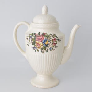 May include: Cream-colored ceramic teapot with a floral design featuring pink, blue, and yellow flowers. The teapot has a lid, a handle, and a spout. The body has vertical ribbed detailing.