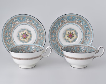 Può includere: Due tazze da tè e piattini bianchi con un motivo floreale decorativo blu e oro. Le tazze da tè hanno un manico e poggiano su un piedino. I piattini hanno un design abbinato con un medaglione floreale centrale.