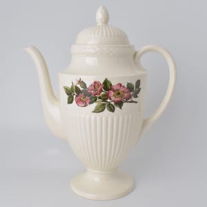May include: Cream-colored ceramic teapot with a lid and handle. The teapot features a floral design of pink roses and green leaves. The body of the teapot has vertical ribbed detailing and sits on a pedestal base. The spout is curved and the handle is a rounded loop.