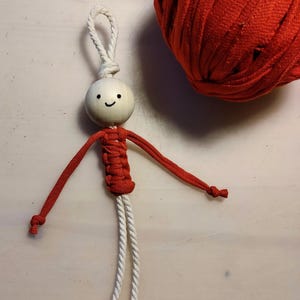 May include: A small, handmade doll with a wooden head and a smiling face. The doll is made with red and white yarn and has long arms and legs.
