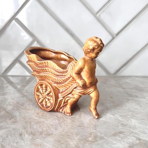 May include: A tan-colored ceramic planter shaped like a chariot being pulled by a cherub. The planter has a detailed design with a wheel and a decorative rim. The cherub is depicted in a walking pose, adding a whimsical touch to the piece.