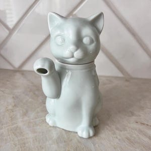 May include: A light blue ceramic cat figurine with a raised paw. The cat has a round head, pointed ears, and a small spout extending from its raised paw. The figurine is sitting upright and has a glossy finish.