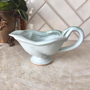 May include: A light blue ceramic gravy boat with a curved handle and a pedestal base. The boat has a wavy rim and a textured interior. The handle and rim have a slightly darker blue-green hue. The gravy boat is on a marble-like surface.