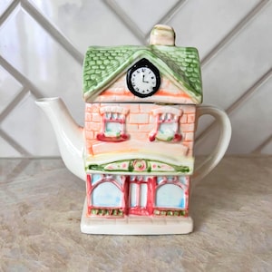 May include: A ceramic teapot shaped like a building, featuring a green roof, a clock, and a brick facade. The teapot has a white spout and handle, with a decorative shop front design. The color palette includes pink, green, and white.