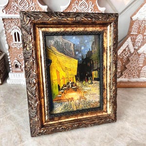 May include: A framed print of Van Gogh's "Cafe Terrace at Night" painting. The artwork depicts a yellow cafe with patrons, set against a dark blue night sky. The frame is ornate, with a dark brown, textured finish.