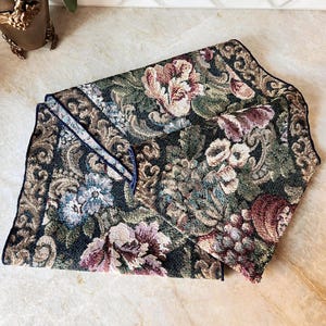 May include: Folded floral tapestry in shades of green, burgundy, and cream. The tapestry features a detailed pattern of flowers and foliage with a dark border. The edges are finished with a dark blue trim.