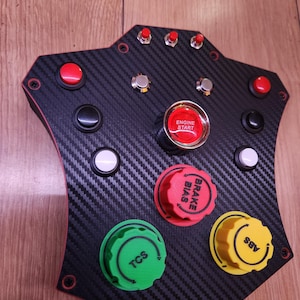 May include: A black carbon fibre control panel with red and silver buttons and knobs. The panel has a red "ENGINE START" button and knobs labelled "BRAKE BIAS", "TCS", and "ABS".