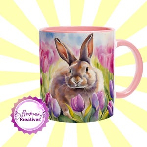 Könnte beinhalten: Eine rosa Keramiktasse mit weißem Innenraum und Henkel. Die Tasse zeigt eine Aquarell-Illustration eines braunen Kaninchens, umgeben von lila Tulpen.
