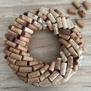 May include: A circular wreath made from numerous used wine corks, displaying various brand names and logos. The corks are arranged in a layered pattern, creating a rustic, natural aesthetic. Some loose corks are scattered around the wreath.
